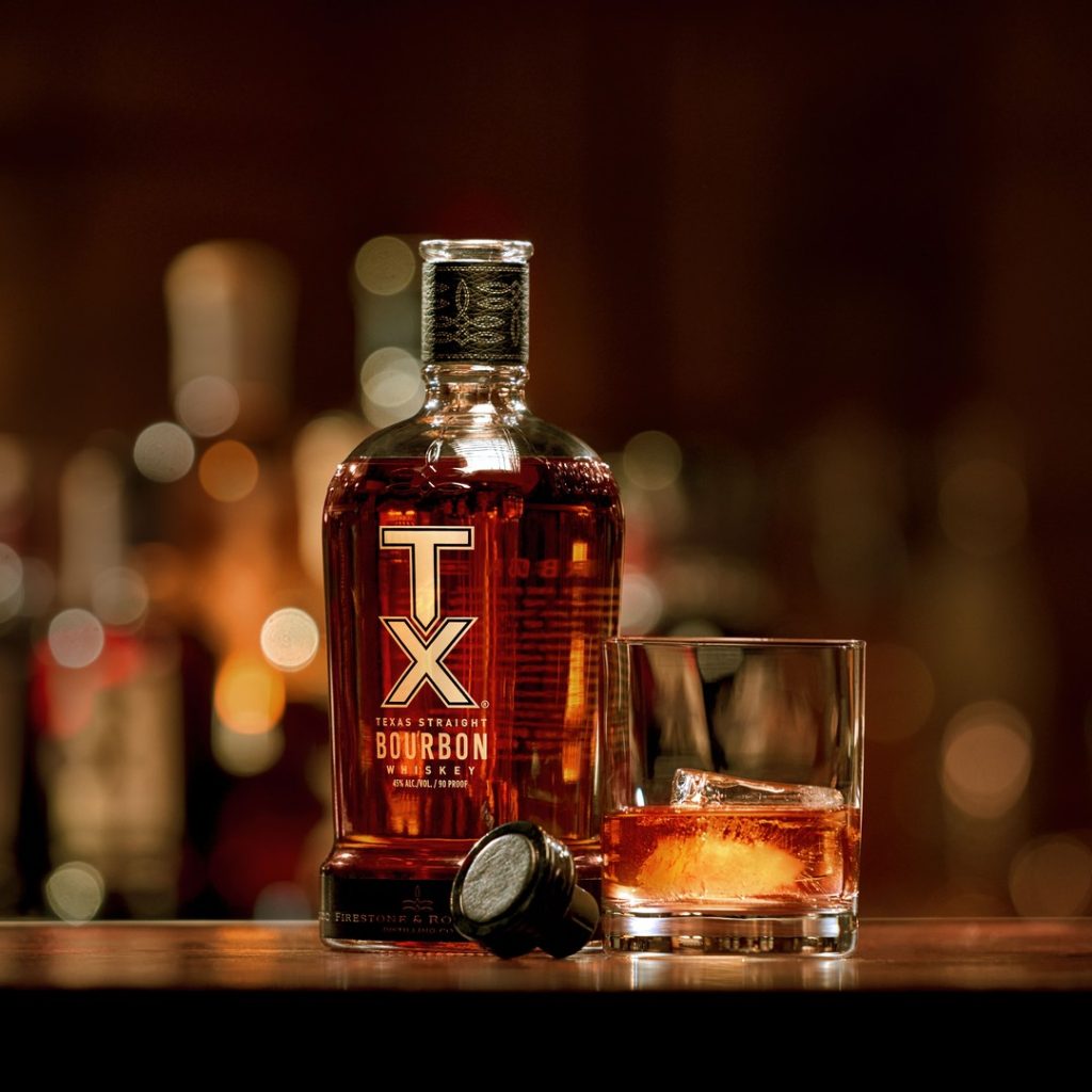 An open TX Whiskey bottle sits on a bar top. Beside it sits its leather cap, along with a rocks glass filled with ice and whiskey. 