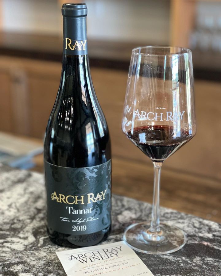 A bottle of Arch Ray 2019 Tannat wine sits on a wooden table next to a glass of red wine branded with the Arch Ray logo. On the table in front of the bottle and glass, you can see the top of a piece of paper with the name "Arch Ray Winery" printed. 