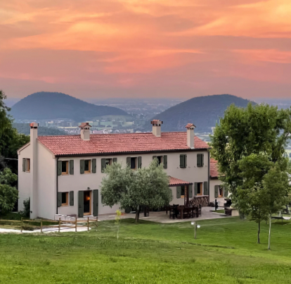 A white house with grey shuttered windows and a terra cotta tile roof sits atop a large green lawn dotted with trees. In the background, there are silhouettes of two large hills woven amongst the sprawling city at their base. The picture is taken at sunset, and the sky is an orange-pink gradient of color. 