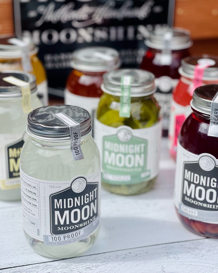 Eight mason jars sitting on a white wooden table filled with Midnight Moon Moonshine of varied flavors. 