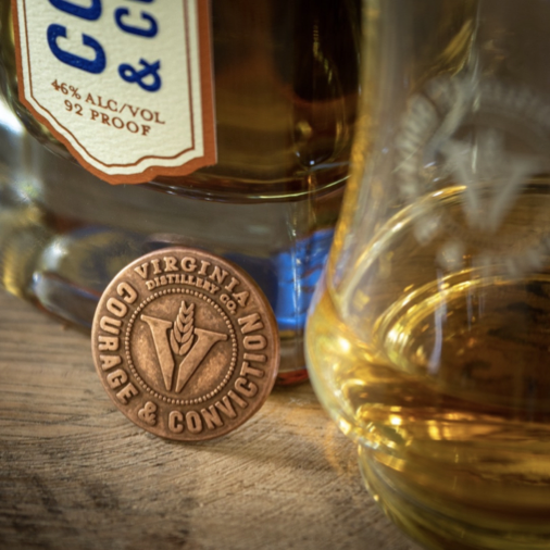 Bottom of a Virginal Distillery Co. bottle with Glencairn glass in the foreground and the VA Distillery magnetic medallion leaning against the bottle. 